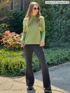 Miik model Johanna (five feet six, xsmall) standing in front of a garden sideway wearing a graphite flare pant along with Miik's Breda funnel neck long sleeve top in green moss and sunglasses 
