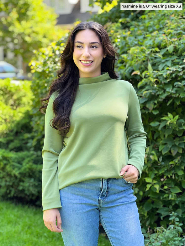 Miik model Yasmine (five feet tall, xsmall, petite) smiling wearing Miik's Breda funnel neck long sleeve top in green moss with jeans 