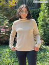 Miik model Meron (five feet three, xsmall) standing in front of a garden wearing Miik's Breda funnel neck long sleeve top in wheat along with a graphite pant