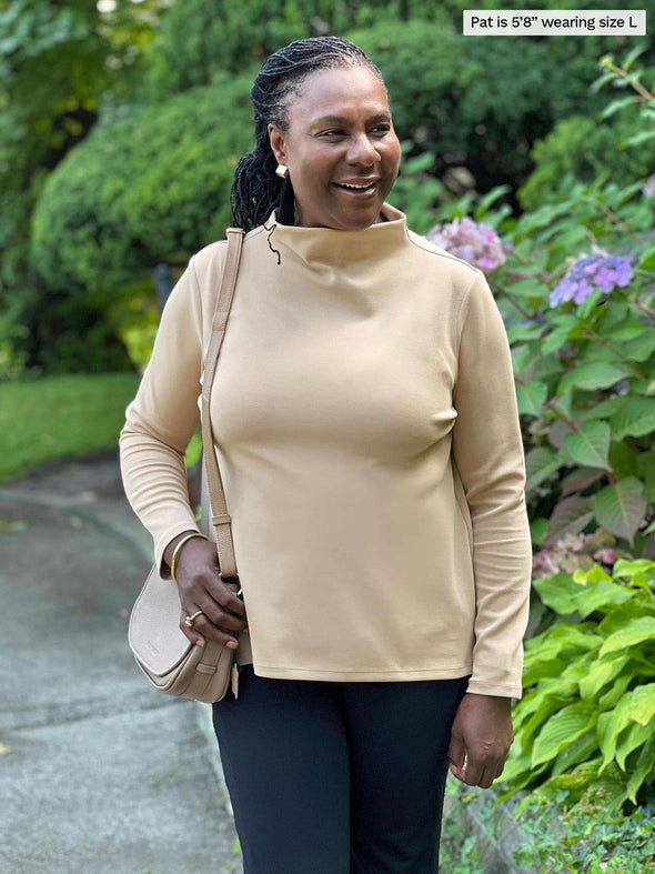 Miik model Pat (five feet eight, large) smiling while looking away wearing Miik's Breda funnel neck long sleeve top in wheat along with a graphite pant and a shoulder bag 