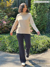 Miik model Meron (five feet three, xsmall) standing in a sidewalk looking away wearing Miik's Breda funnel neck long sleeve top in wheat with a graphite flare pant 