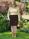 Miik model Bri (five feet five, xlarge) standing in a garden while looking away wearing Miik's Breda funnel neck long sleeve top in wheat with a pencil skirt in dark chocolate  