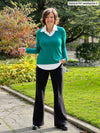 Miik founder Donna (5'6", medium) standing on a sidewalk smiling wearing Miik's Carson collared faux-layer shirt in jade melange/white with a wide leg pant in black and boots 