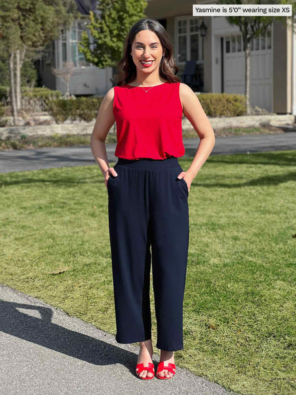 Miik model Yasmine (5'0", xsmall, petite) smiling with both hands on pockets wearing Miik's Cassandra pull-on pocket capri pant in navy with a poppy red top 