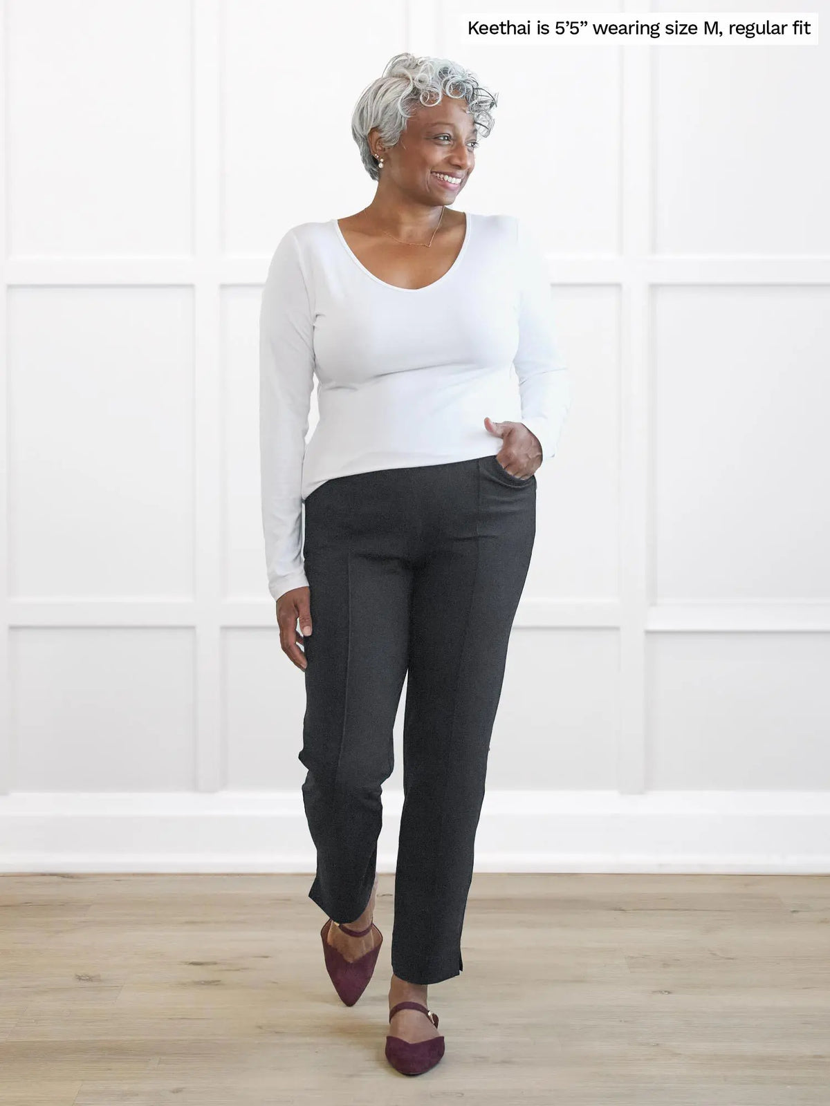 Woman wearing a white long-sleeve top and dark pants standing against a white wall 