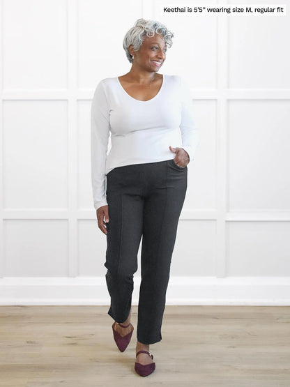 Woman wearing a white long-sleeve top and dark pants standing against a white wall 