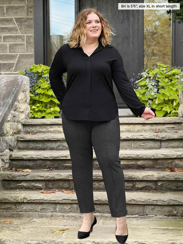 Miik model Bri (5'5", xlarge) smiling and looking up wearing Miik's Christal pull-on pintuck ankle pant in charcoal short length along with a collared black shirt