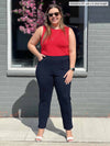 Miik model Christal (5'3", large) standing in front of a window/brick wall wearing a poppy red tank along with Miik's Christal pull-on pintuck ankle pant in navy short length