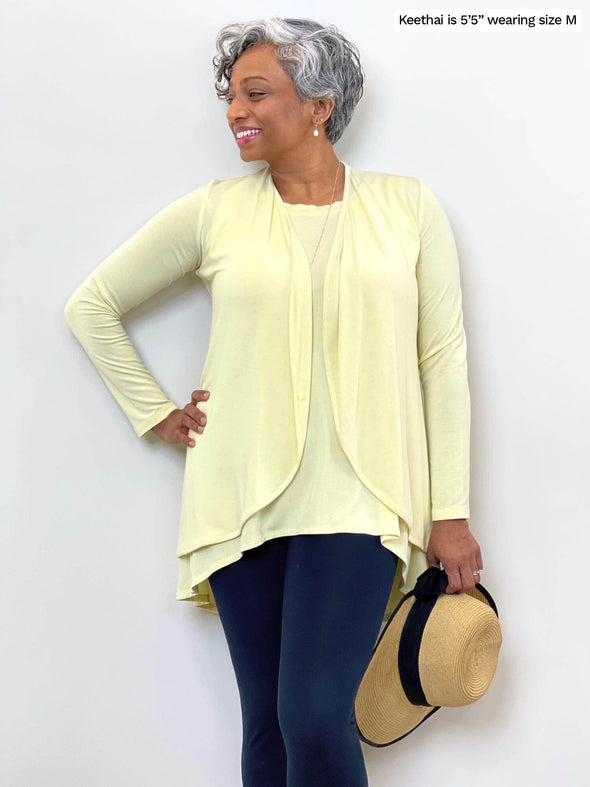 Woman standing in front of a wall wearing Miik's Dalya high-low flowy tunic tank in yellow with matching cardigan and navy pants.