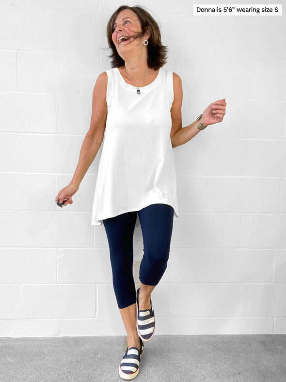 Woman standing in front of a wall wearing Miik's Dalya high-low flowy tunic tank in white with navy leggings.
