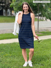 Miik model Johanna (five feet six, xsmall) standing outside and smiling wearing Miik's Dani high neck sleeveless dress in navy wide pinstripe holding a white denim jacket over her shoulders 