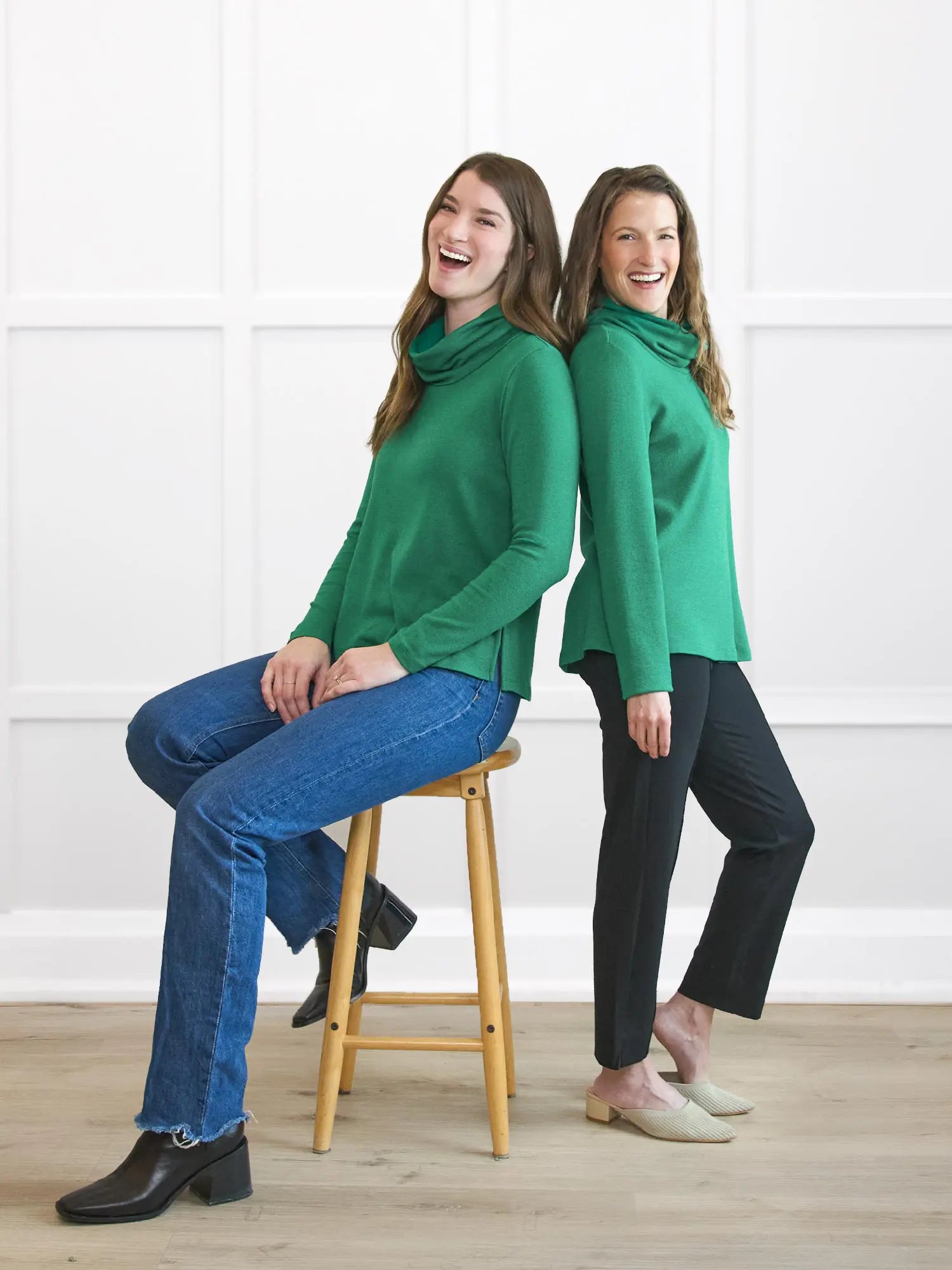 Two women wearing green cowl-neck tops in a room with a white wall and wooden floor 