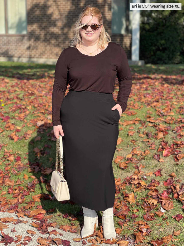 Woman standing in nature wearing Miik's Devon pocket midi skirt in black with a brown top.