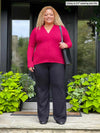 Miik model Carley (5'2", xxlarge) standing in a entry way smiling wearing a black pant along with Miik's Dua v-neck long sleeve blouse in bordeaux