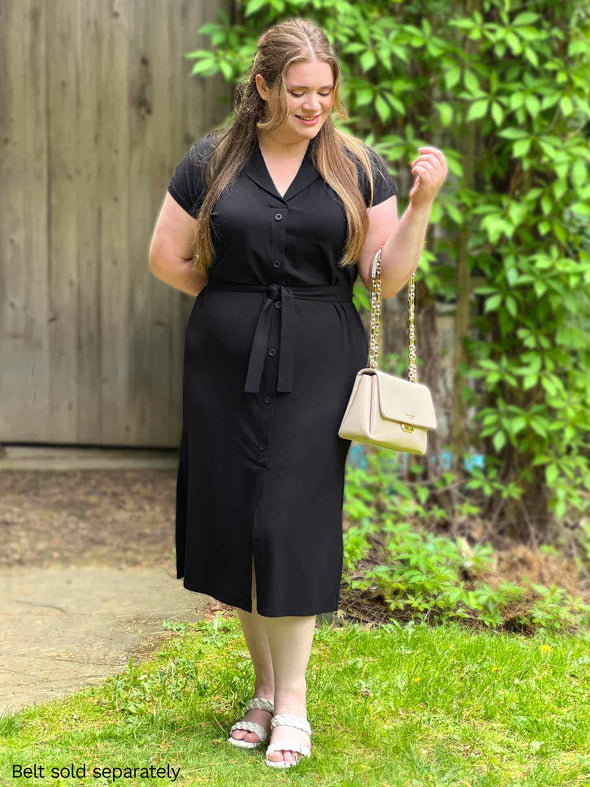 Woman standing in nature wearing Miik's Dylan collared midi sheath dress in black with the blair belt in black colour while holding a cream colour purse and looking down.