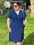 A woman wearing a navy blue wrap-style dress with a side twist detail, standing outdoors on a green lawn. She is wearing round sunglasses, a gold necklace, and carrying a small silver and black patterned shoulder bag. 