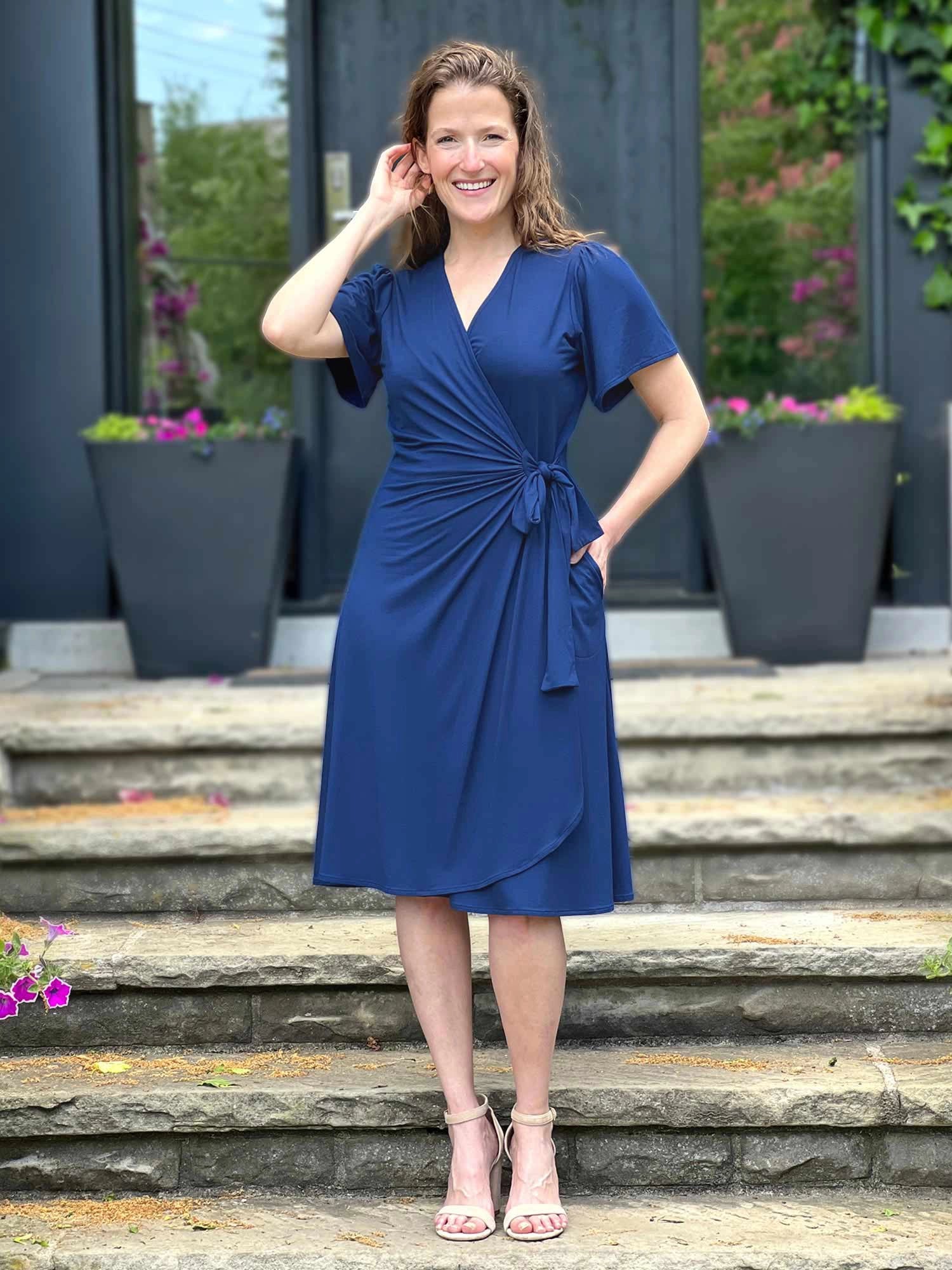 A woman wearing a royal blue wrap-style midi dress with short flutter sleeves and a twisted waist detail, standing on stone steps in front of a modern building entrance. 
