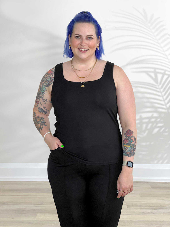 Miik model Kaitlin (5'9", xlarge) smiling while standing in front of a white wall wearing Miik's Eline reversible shelf bra tank in black along with a pant in the same colour 