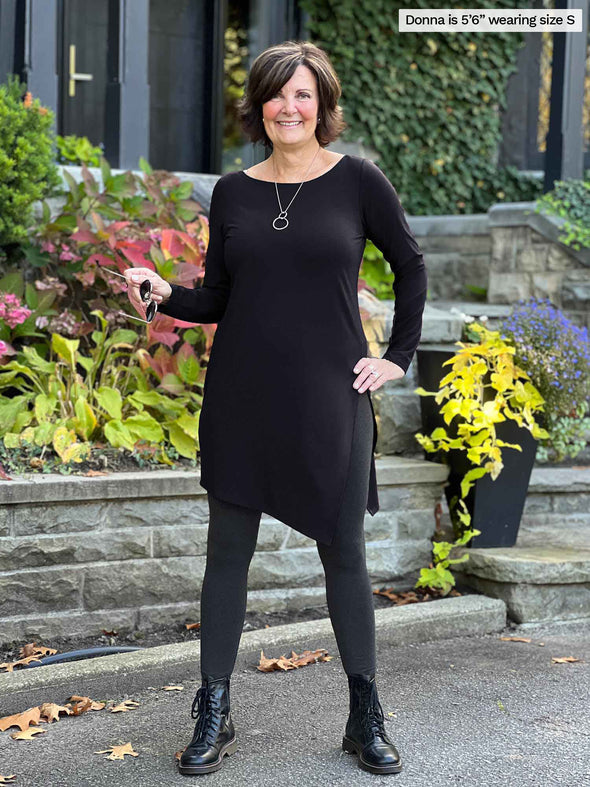 Miik founder Donna (5'6", small) smiling while stand on a front door wearing Miik's Elsie asymmetrical long sleeve tunic in black with a charcoal legging and boots 