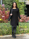 Miik model Yasmine (5'0", xsmall, petite) smiling holding a purse wearing Miik's Elsie asymmetrical long sleeve tunic in black with a charcoal legging and black boots 