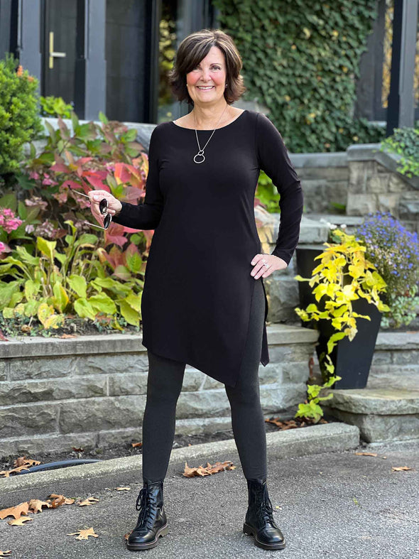 Miik founder Donna (5'6", small) smiling while stand on a front door wearing Miik's Elsie asymmetrical long sleeve tunic in black with a charcoal legging and boots