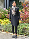 Miik model Meron (5'3", xsmall) smiling and looking away wearing a black legging along with Miik's Elsie asymmetrical long sleeve tunic in charcoal and black loafers 