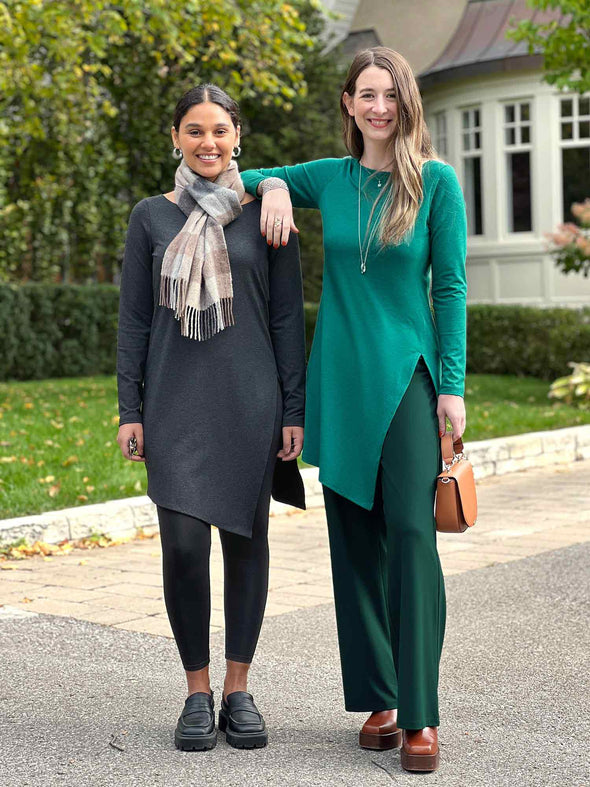 Miik models Meron and Johanna (both xsmall) smiling while standing next to each other wearing Miik's Elsie asymmetrical long sleeve tunic in charcoal, Meron, and jade melange, Johanna
