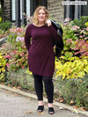 Woman standing in nature wearing Miik's Elsie asymmetrical long sleeve tunic in port with leggings.