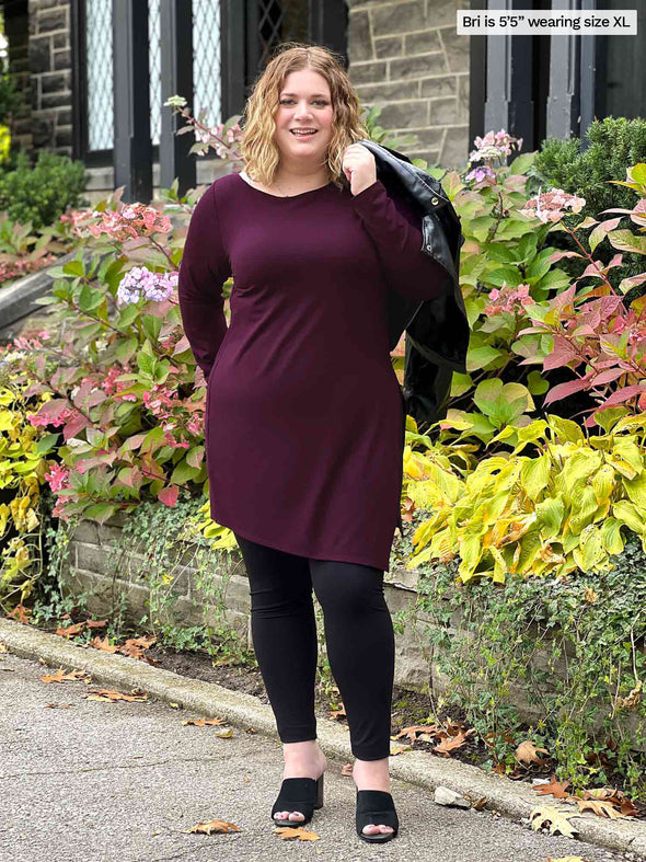 Woman standing in nature wearing Miik's Elsie asymmetrical long sleeve tunic in port with leggings.