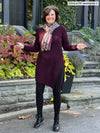 Miik founder Donna (5'6", small) smiling wearing Miik's Elsie asymmetrical long sleeve tunic in port along with black legging and a printed scarf, boots and sunglasses 