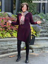 Woman standing in front of a house wearing Miik's Elsie asymmetrical long sleeve tunic in port with leggings.