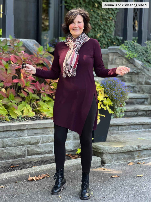 Miik founder Donna (5'6", small) smiling wearing Miik's Elsie asymmetrical long sleeve tunic in port along with black legging and a printed scarf, boots and sunglasses 