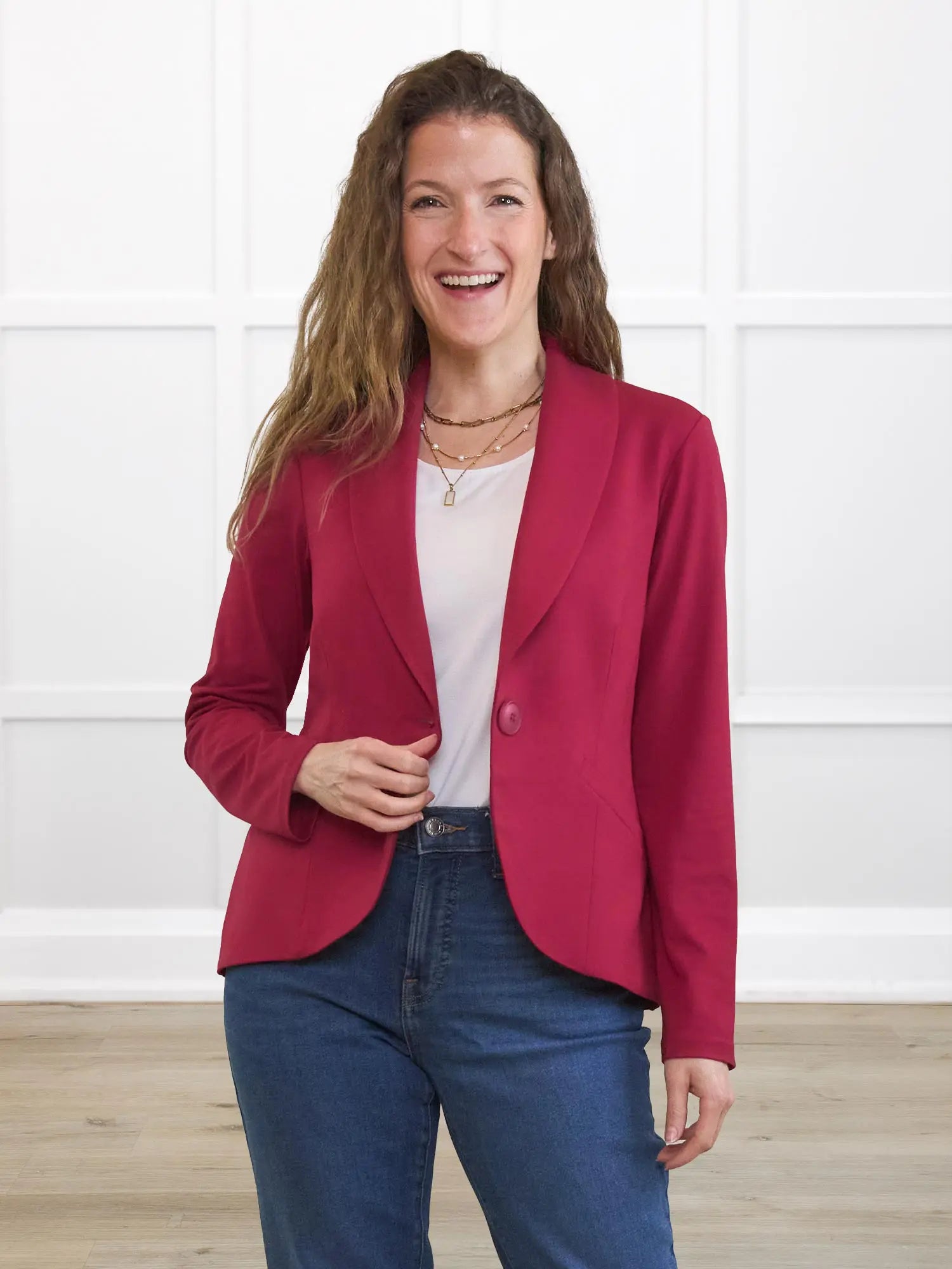 Woman wearing a red blazer over a white shirt and blue jeans, standing against a white wall.