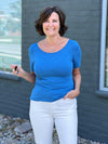 Miik founder Donna (five feet six, small) smiling in front of a brick wall wearing Miik's Emmy reversible half sleeve top in cobalt melange with a white jeans