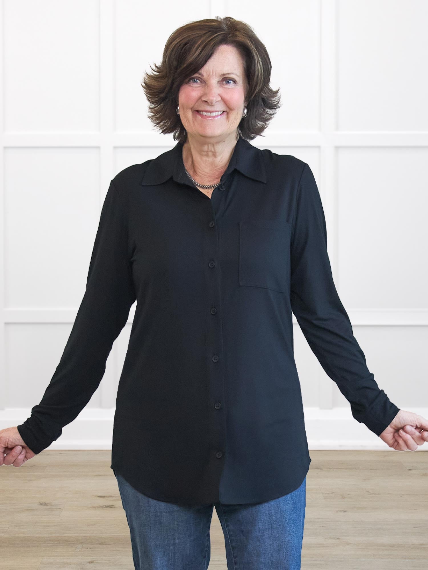A smiling woman with short brown hair wearing a black long-sleeve button-up shirt and blue jeans, standing against a white paneled wall with light wood flooring.