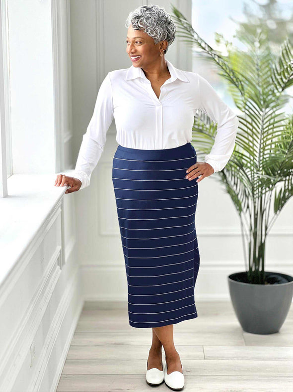 Miik model Keethai (five feet five, size medium) standing next to a wall looking away wearing a collared white shirt along with Miik's Frankie midi skirt in navy wide pinstripe and white shoes