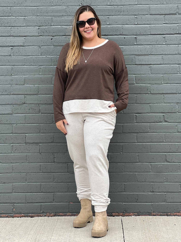 Woman standing in front of a brick wall wearing Miik's Geneva two-tone crew neck top in brown chocolate melange/oatmeal, oatmeal fleece jogger, boots and sunglasses
