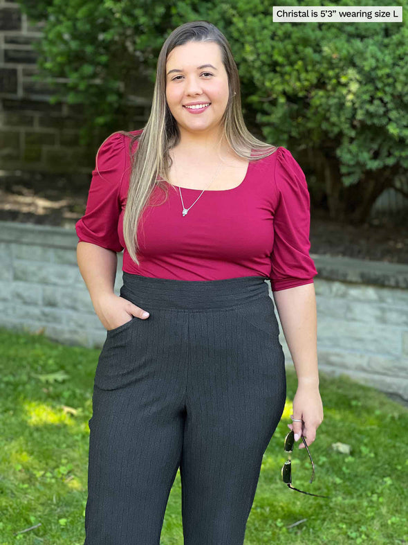 Miik model Christal (five feet three, large) smiling wearing Miik's Gigi reversible puff sleeve blouse in bordeaux tucked in a pinstripe charcoal pant