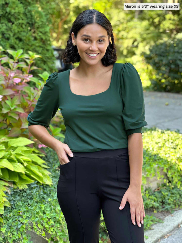 Woman standing in nature wearing Miik's Gigi reversible puff sleeve blouse in pine green with black pants.