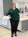 Miik plus size model Jennifer (5'2", 4x) smiling while standing on a living room wearing Miik's Gracelyn v-neck classic tee in pine green with a black pant 