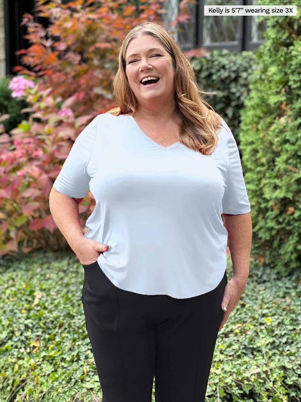 Miik plus size Kelly (5'7", 3x) laughing with one hand on the pant pocket wearing Miik's Gracelyn v-neck classic tee in white with a dress black pant 