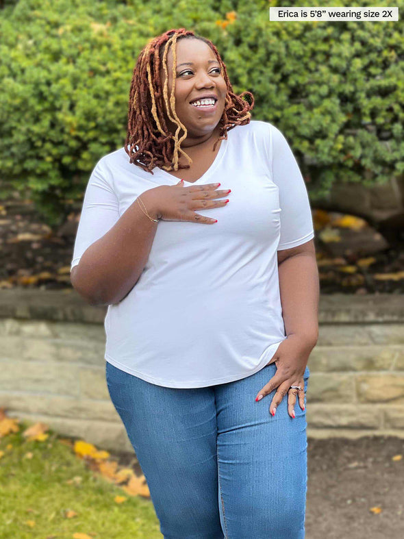 Miik plus size model Erica (5'8", 2x) smiling and looking up with on hand on her chest looking surprised wearing Miik's Gracelyn v-neck classic tee in white and jeans