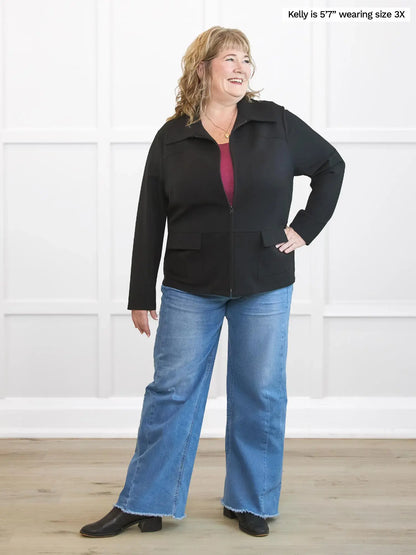 Woman wearing a black jacket and blue jeans standing against a white wall. 