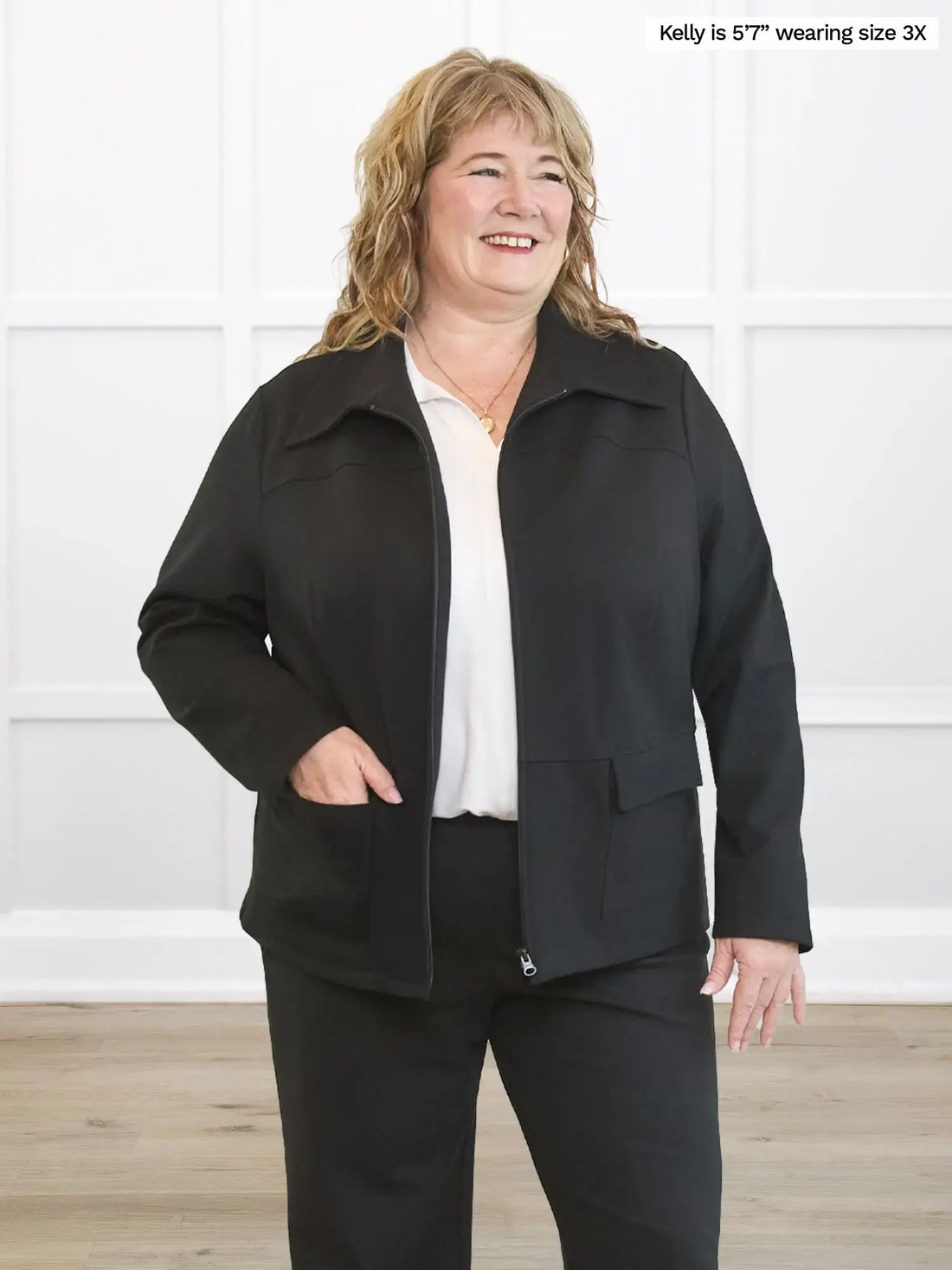 Woman wearing a black coat and pants against a white wall. 