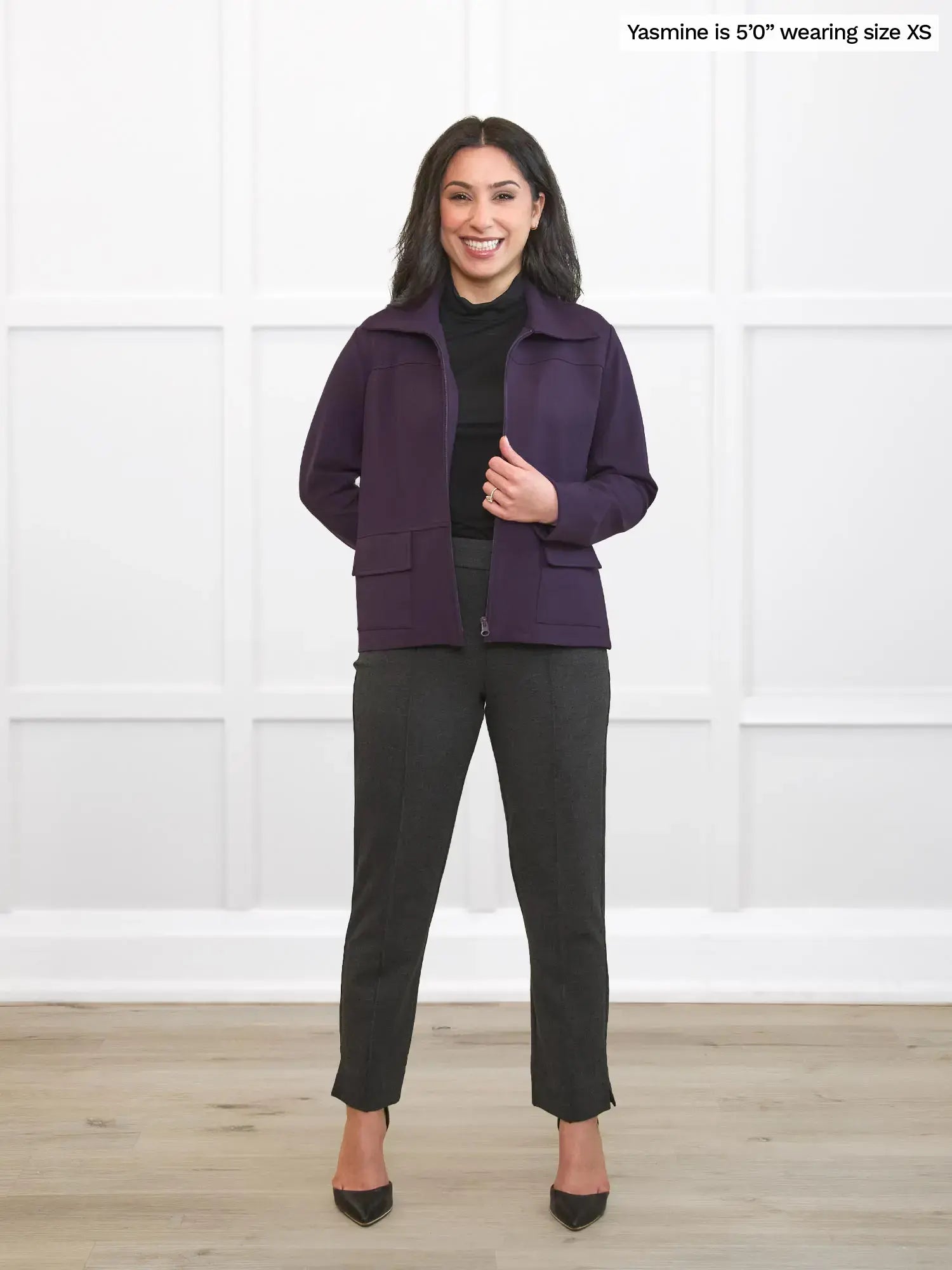 Woman wearing a purple jacket and black pants standing against a white wall. 