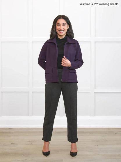 Woman wearing a purple jacket and black pants standing against a white wall. 