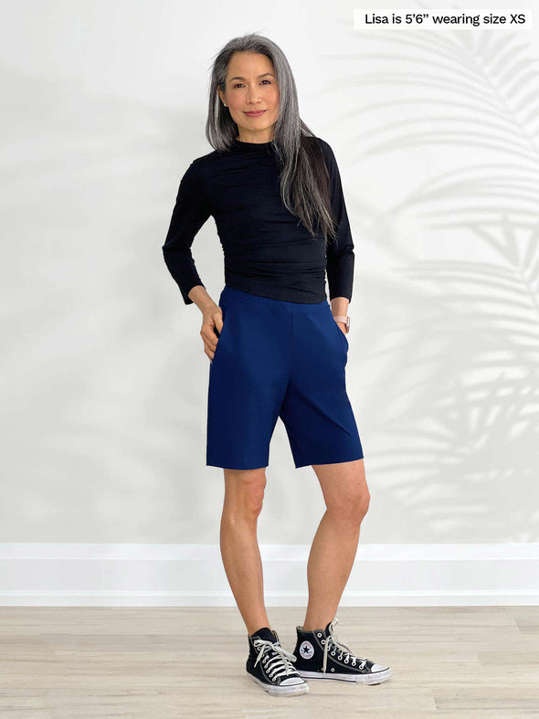 Miik model Lisa (5'6", xsmall) standing in front of a white wall wearing Miik's Irelynn bermuda pocket short in ink blue with a long sleeve top in black 