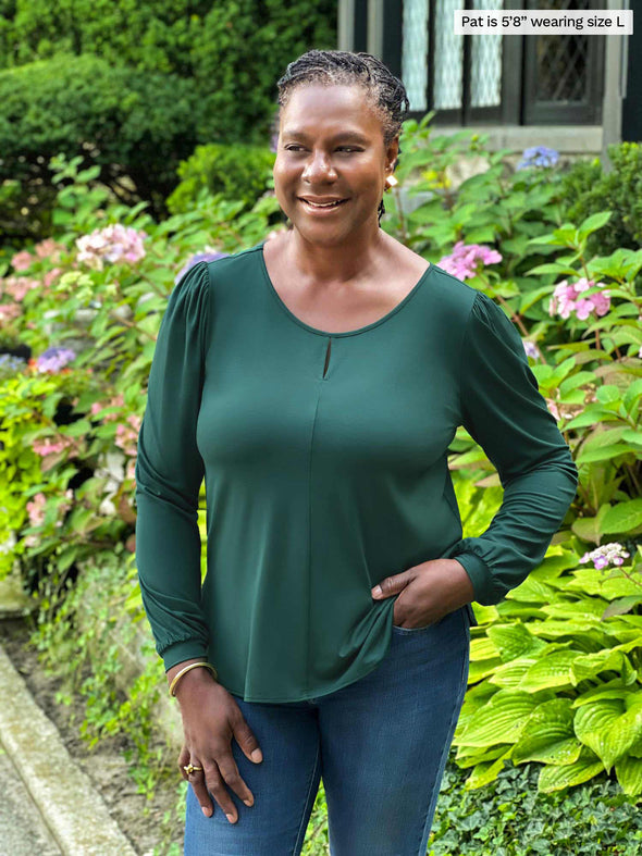 Miik model Pat (5'8", large) smiling and looking away wearing Miik's Janette puff sleeve reversible blouse in pine green with jeans 