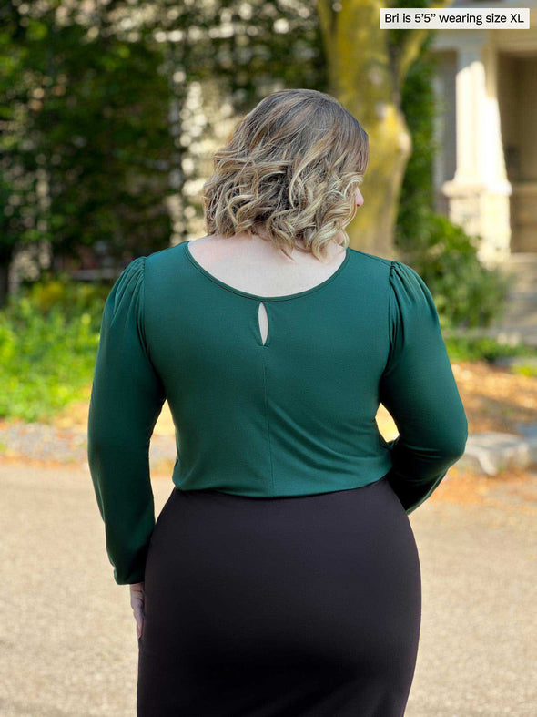 Miik model Bri (5’5”, xlarge) standing with her back towards the camera showing the back of Miik's Janette puff sleeve reversible blouse in pine green showing the blouse reversed 