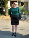 Miik model Bri (5'5", xlarge) looking away awhile smiling wearing Miik's Janette puff sleeve reversible blouse in pine green with a pencil skirt in black and boots 